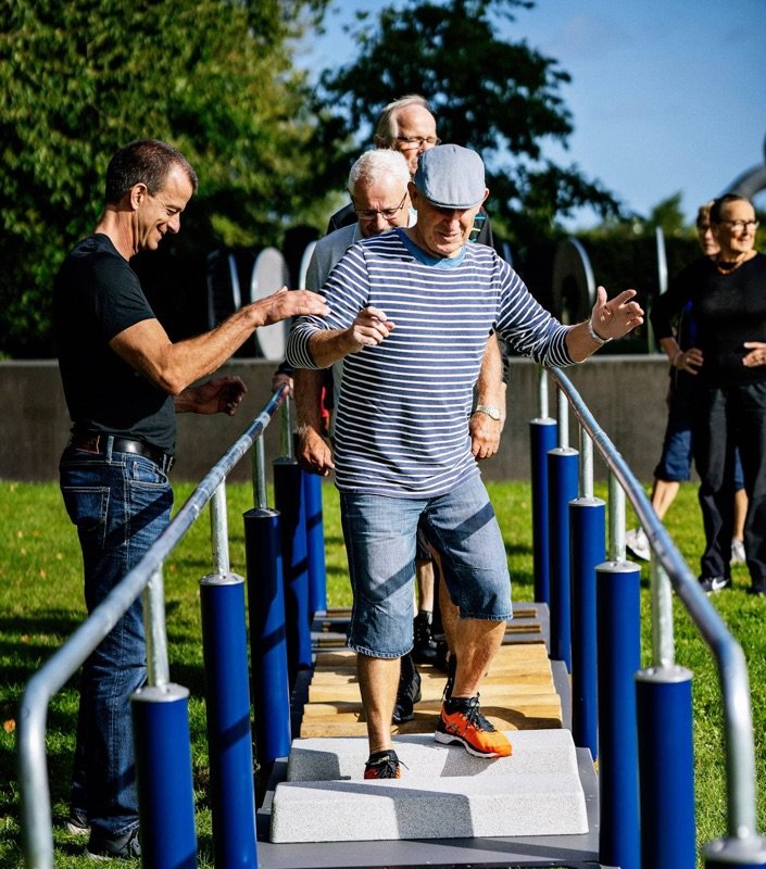 En gruppe ældre træner balance på en udendørs forhindringsbane, mens en instruktør støtter manden forrest.