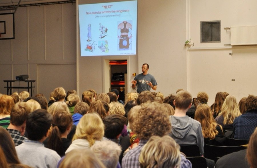 Undervisning foran en tætpakket sal med en slide om NEAT — non-exercise activity thermogenesis — på lærredet.